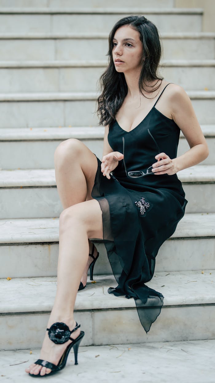 Chic woman in a black dress sitting thoughtfully on marble stairs holding glasses.